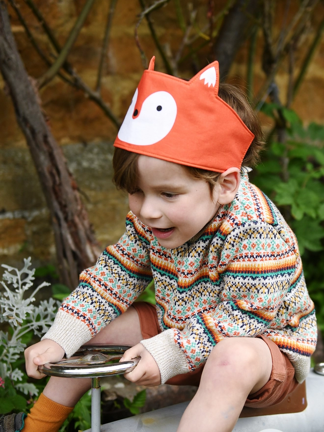 A young child wearing a knitted jumper and a fox dress up fabric crown sits on a ride on vintage car in the garden.