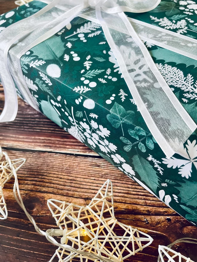 Close-up of present wrapped in emerald green festive leaf wrapping paper with Holly and Ivy design. Star lights surround on wood background