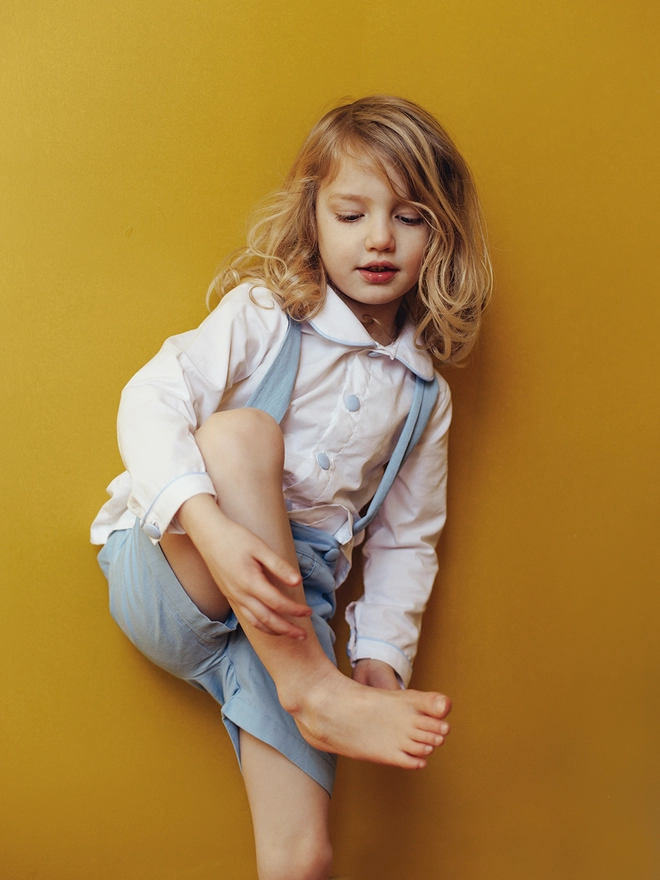 A little boy stands against a yellow wall holding one foot up  wearing a white peter pan  collared shirt and blue shorts with braces