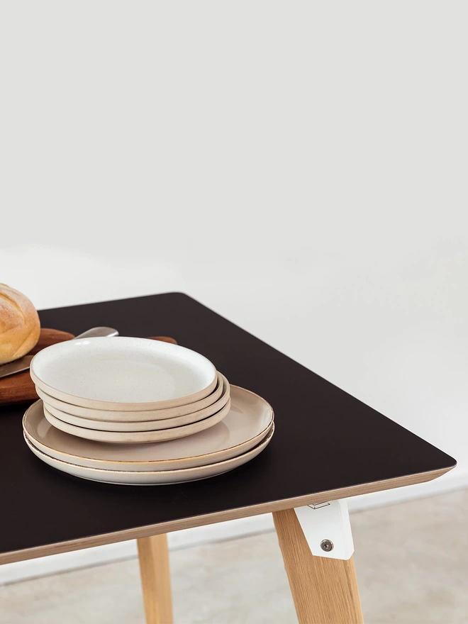 Corner view of a pile of plates on a stylish and minimalist dining table with Fenix top, coloured steel brackets and solid oak legs.