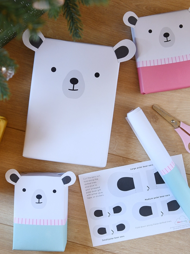 Three gifts wrapped as polar bears lay on a wooden floor beside a sheet of wrapping paper and scissors.