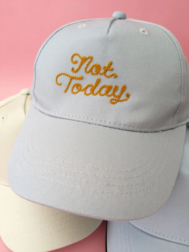 A stack of kids baseball caps in Powder Blue & Pistachio Green. The blue hat has Gold embroidery reading "Not Today" on a pink background