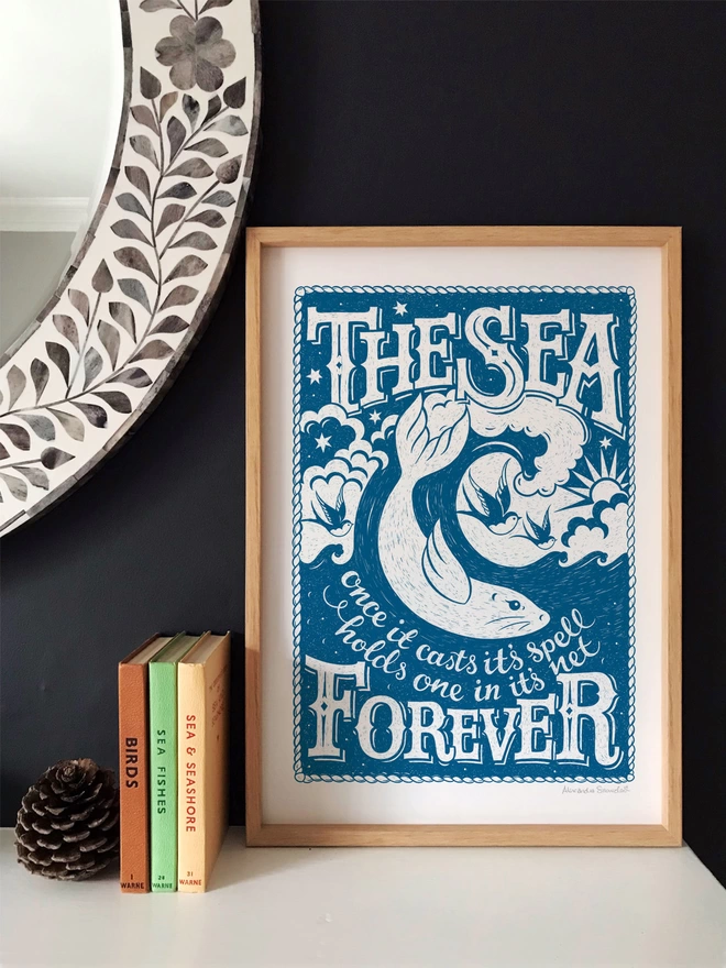 Blue sea print with books, fir cone and mirror against black wall