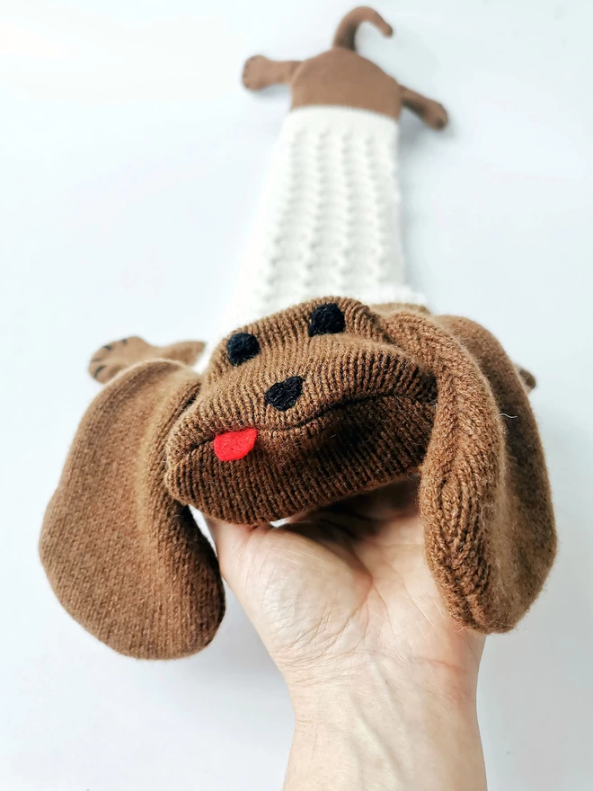 A full length image of Dolly the sausage dog long hot water bottle showing her hand embroidered face, floppy ears and cream cable knit coat. A hand tickles her under the chin like a real dog.