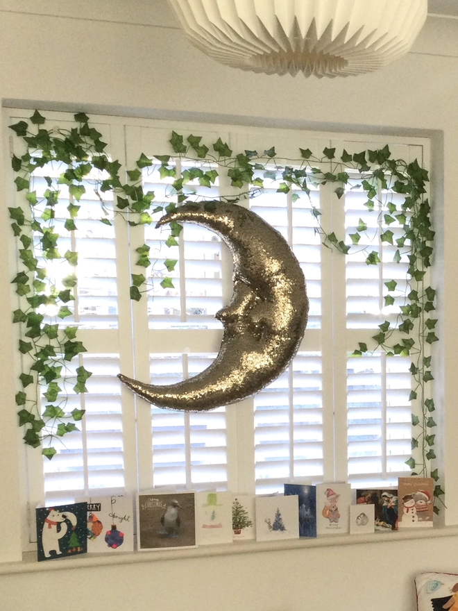 A large gold sequin plush half moon decoration, with faces, hangs in front of a white shuttered window surounded by ivy and Christmas cards
