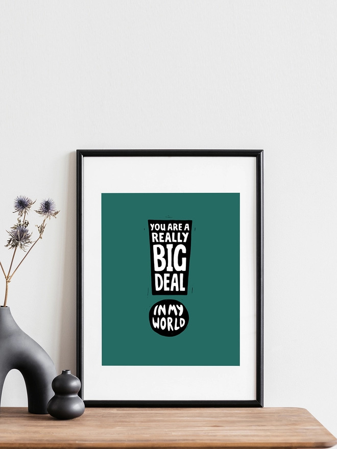 A Woodism print in a black frame propped on a shelf in a stylish home. The print is green with an exclamation mark design made from typographic words which say: You Are A Really Big Deal In My World.