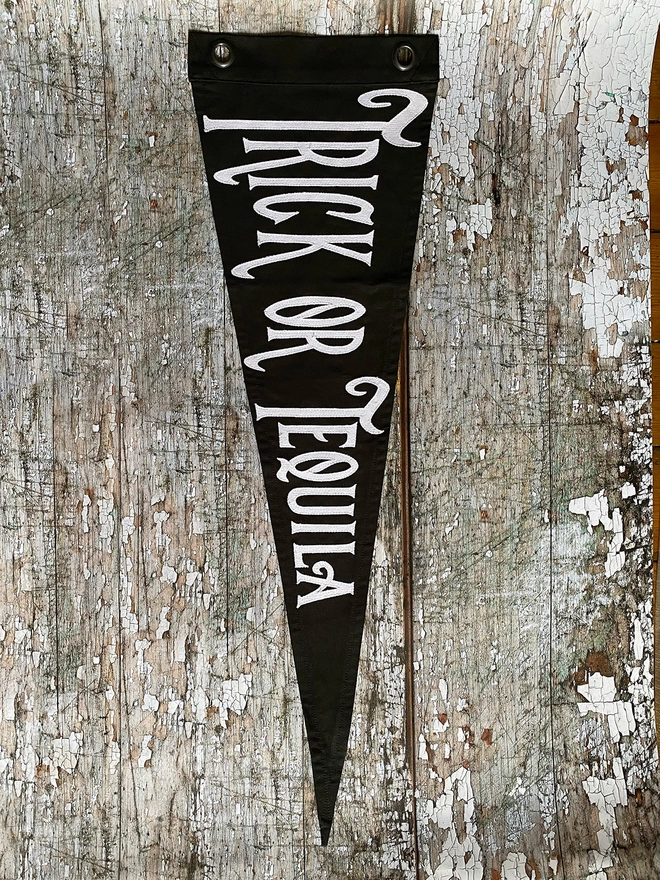 A black pennant flag hanging vertically on a wooden wall. In white canvas letters is written Trick or Tequila