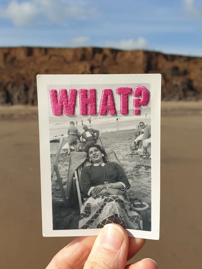 B & W photo of woman with pink embroidered ‘What?”’ held against beach background 