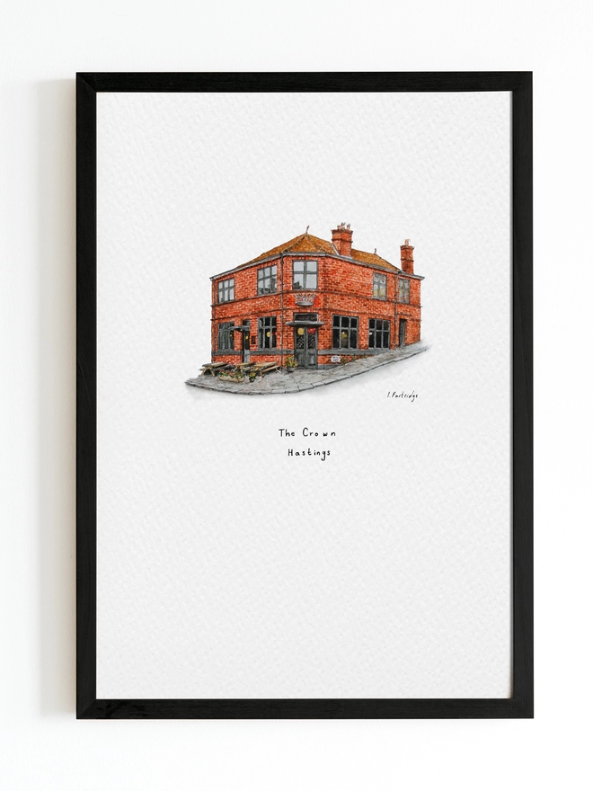 Watercolour illustration of The Crown in Hastings a beautiful brick fronted pub with a characterful appearance and a metalwork crown sitting above the entrance door. The illustration is a small watercolour painting in the centre of the white page and sits within a black frame. 