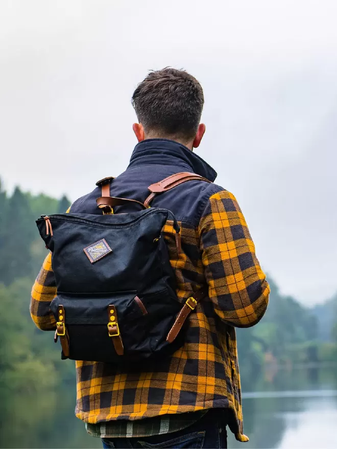 Model standing by lake wearing black zip top backpack with yellow hardware.