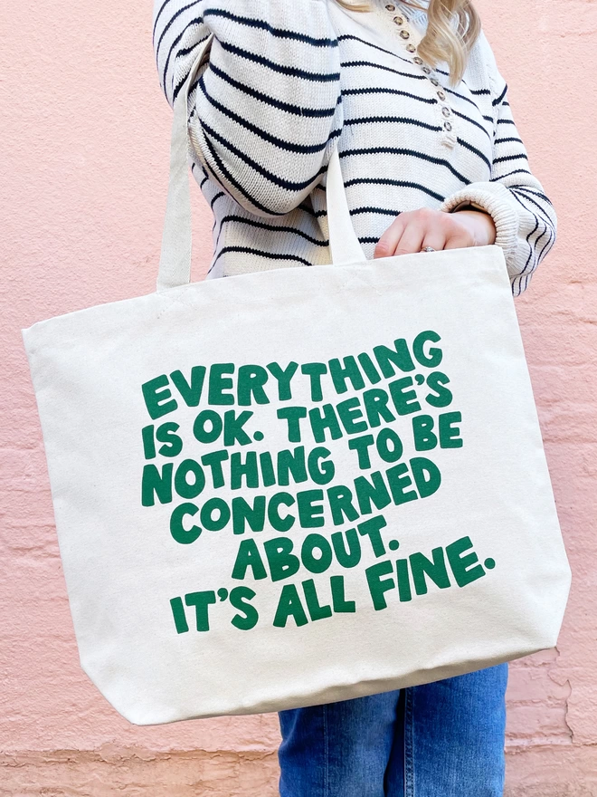 model holding a large natural canvas tote bag bearing the words Everything is OK. There's nothing to be concerned about. It's all fine.