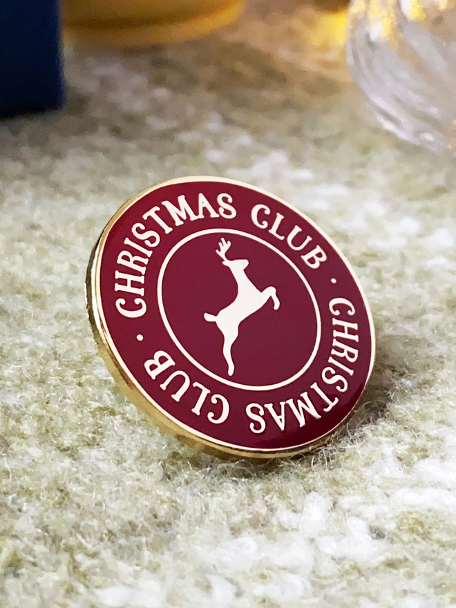A deep red and gold enamel pin badge with a gold reindeer in the centre and the words "Christmas Club" around the outside is on a green blanket.