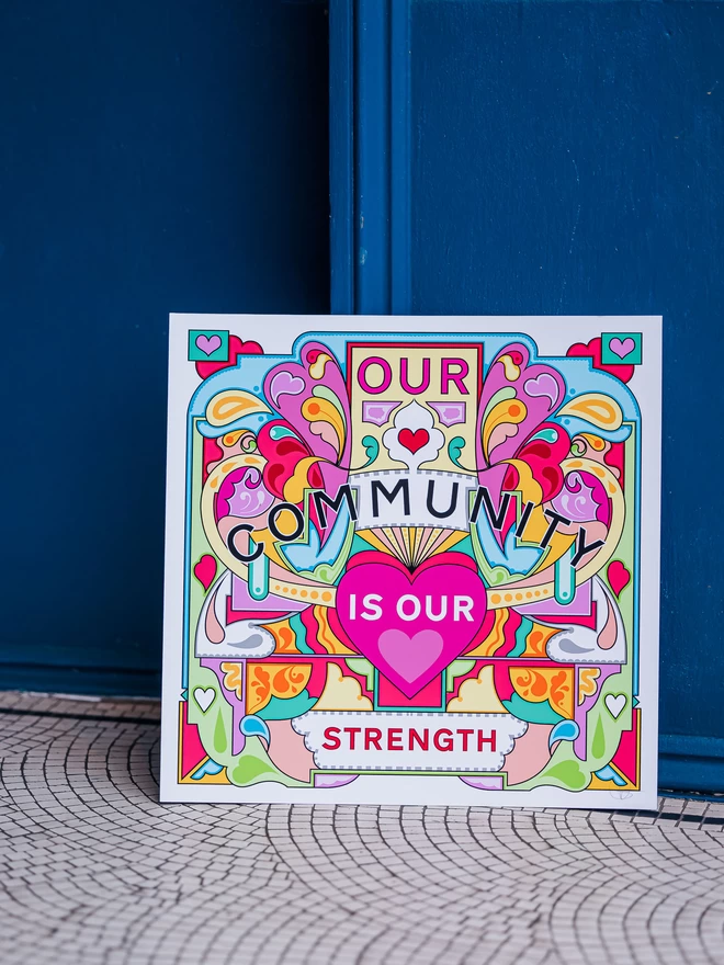 Our Community is Our Strength is written over this bold, symmetrical illustration of yellows, greens and pinks. The print sits on a tiled white floor and is resting against a dark blue wall. 