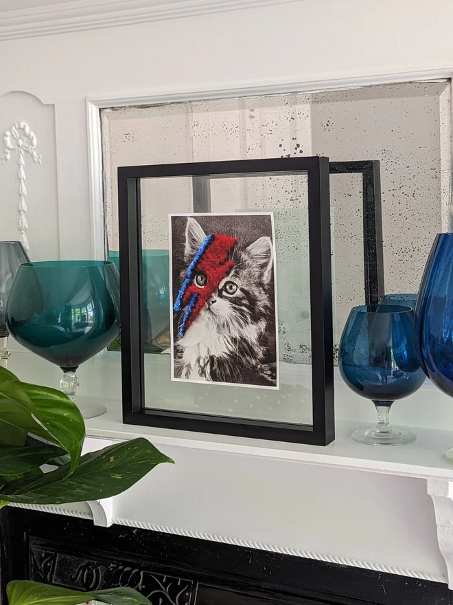 Black and white cat print with red and blue embroidered bolt in frame held on fireplace
