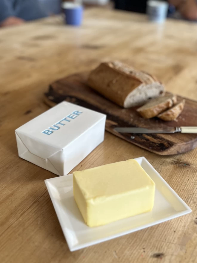 A block of butter sits on a handmade ceramic dish. The top of the lid has ‘butter’ painted in blue.