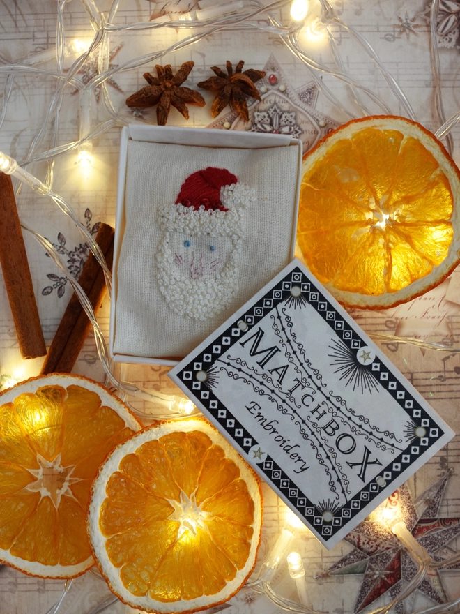 An embroidered Santa displayed in its matchbox, surrounded by fairy lights, dried orange slices, cinnamon sticks and star anise.