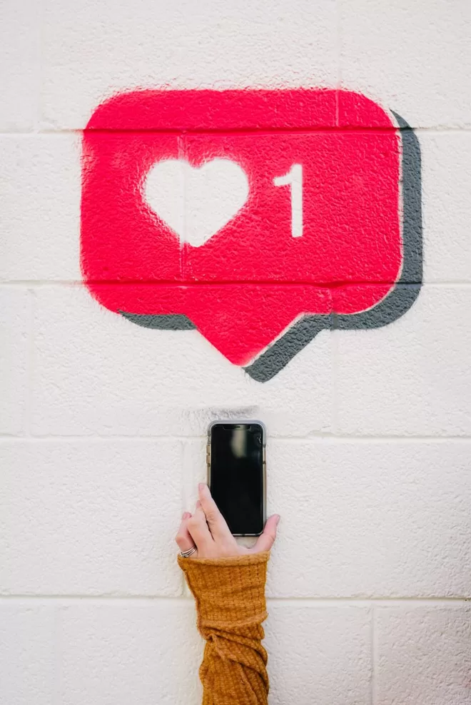 Instagram Like symbol painted on a white wall with someone taking a picture of it with a mobile phone. 