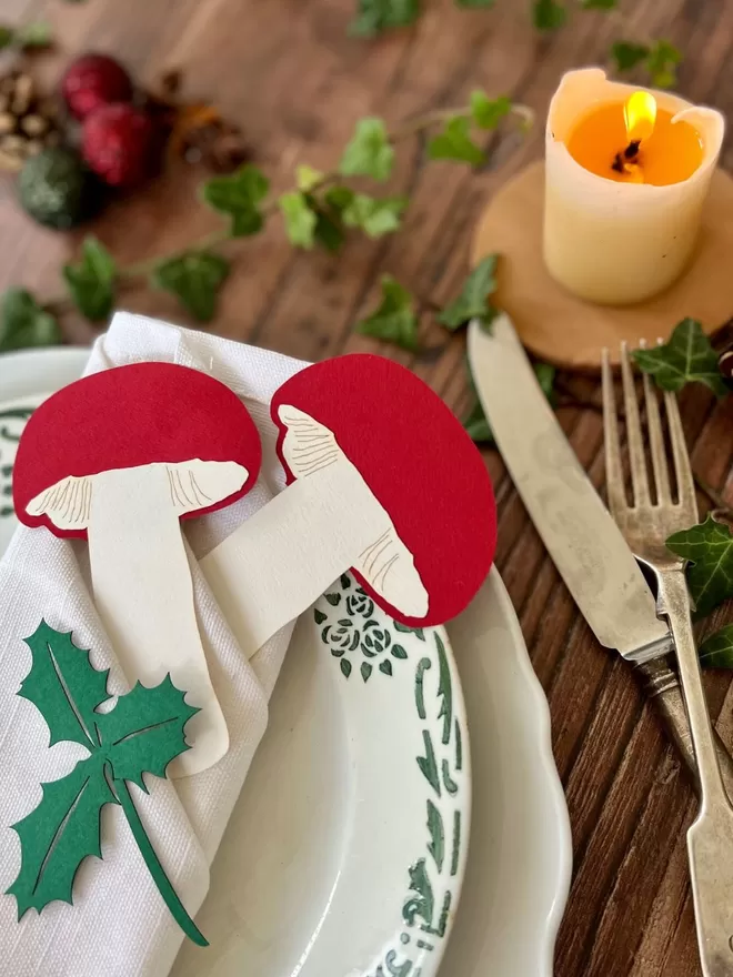Christmas Place Setting showing two red and white paper toadstools and a paper holly sprig placed on a white folded napkin