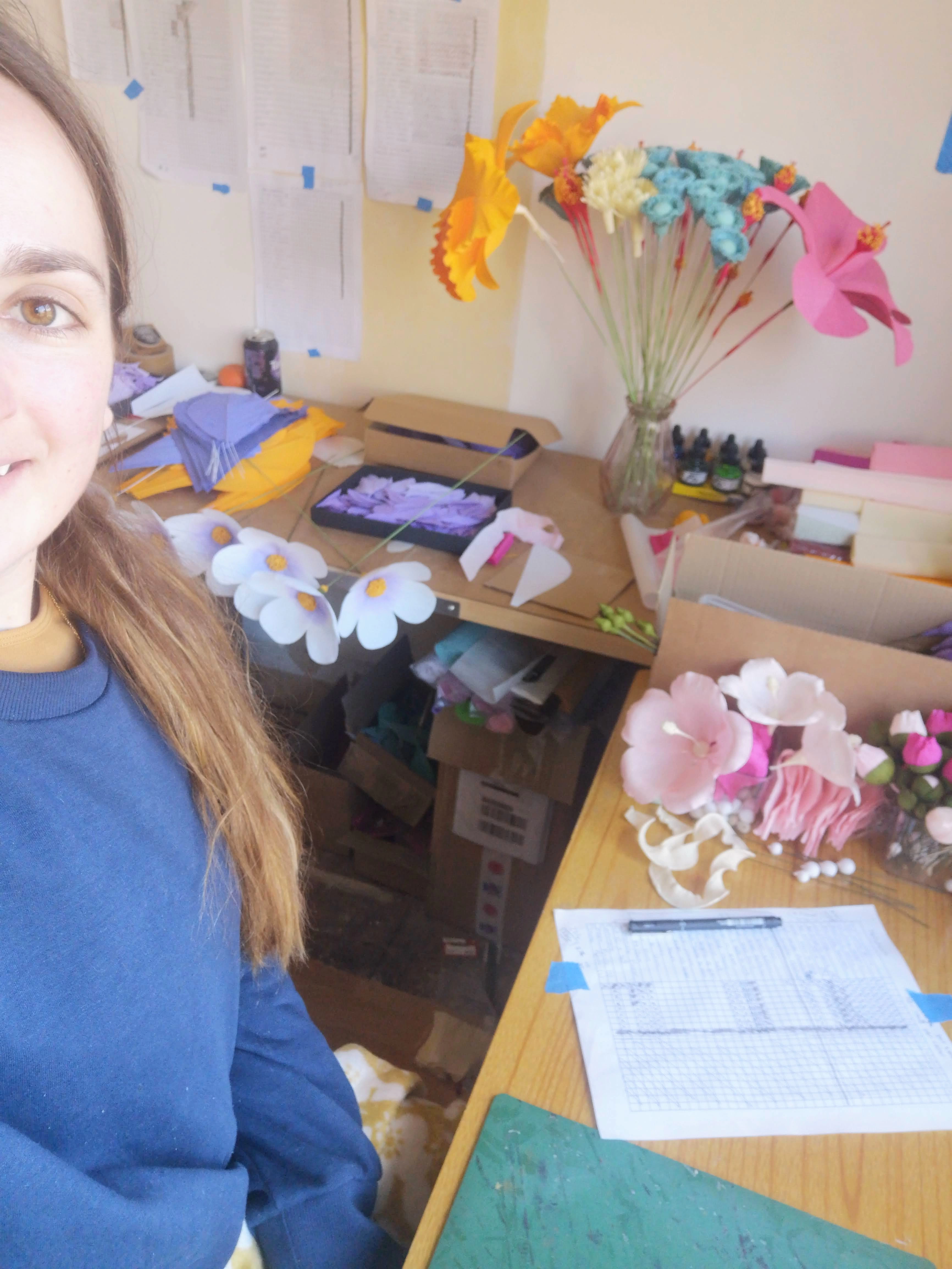 Artist photographed sitting at a desk, surrounded by paper flowers and to do lists