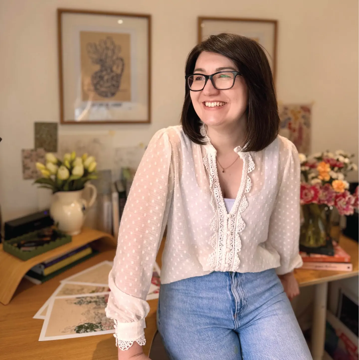 Jenna Crush sitting on studio desk in front of art prints on desk and wall