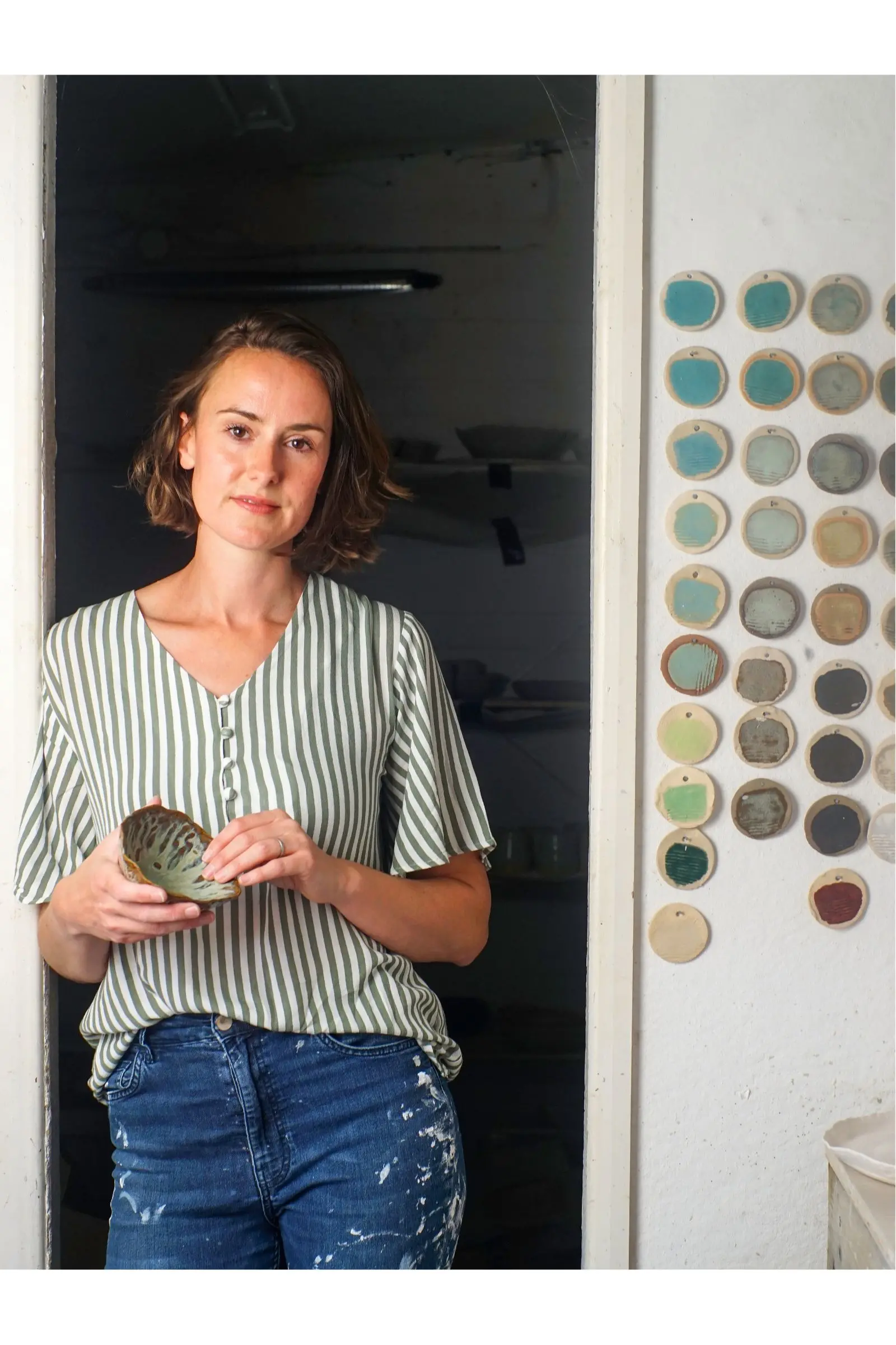 Jenny Hopps J.Hopps Pottery standing in the doorway of her studio wearing a striped shirt. There are ceramic samples on the wall and Jenny is holding a ceramic bowl.