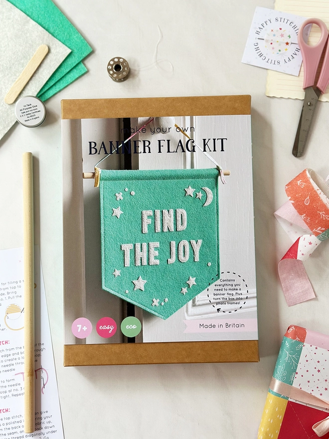 A craft kit box to make a felt positive message banner lays on a white desk with the components surrounding the box.