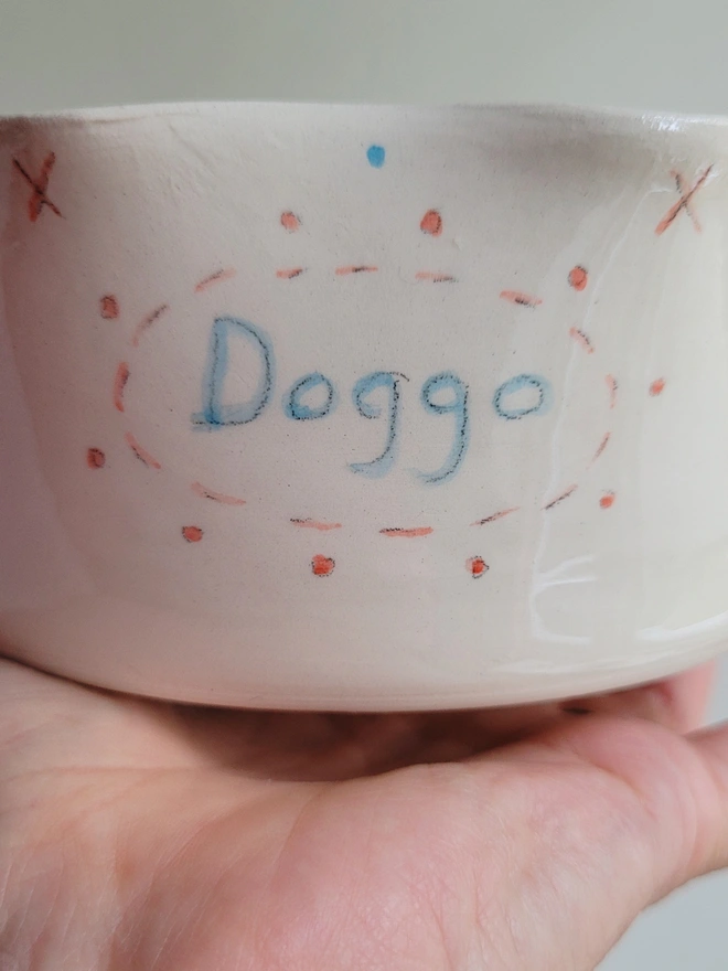 pottery dog bowl close up showing the hand painted word doggo in blue with red dots surrounding it