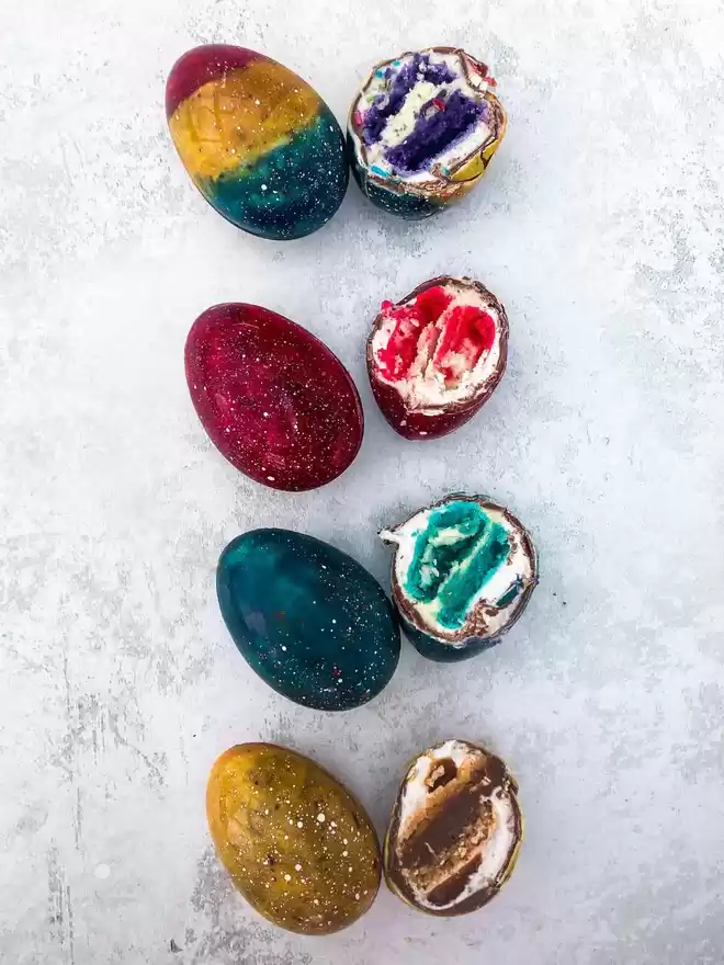a group of colourful easter eggs on top of a white surface some cut open showing a marshmallow and macaron centre