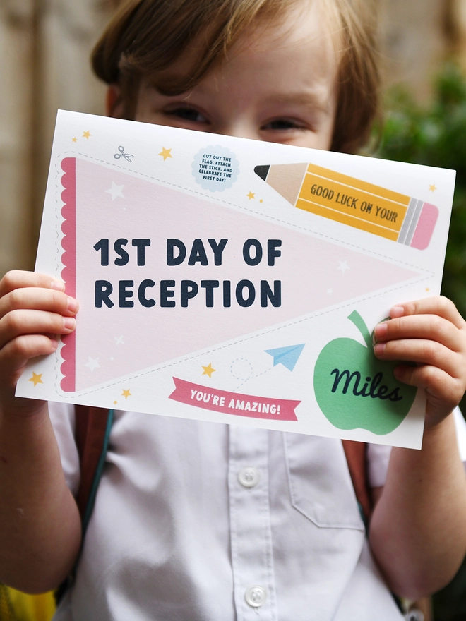 A young child, wearing a school uniform and backpack, holds a "1st Day of Reception" card with excitement. The card features encouraging messages and playful designs, perfect for celebrating a child's first day at school.