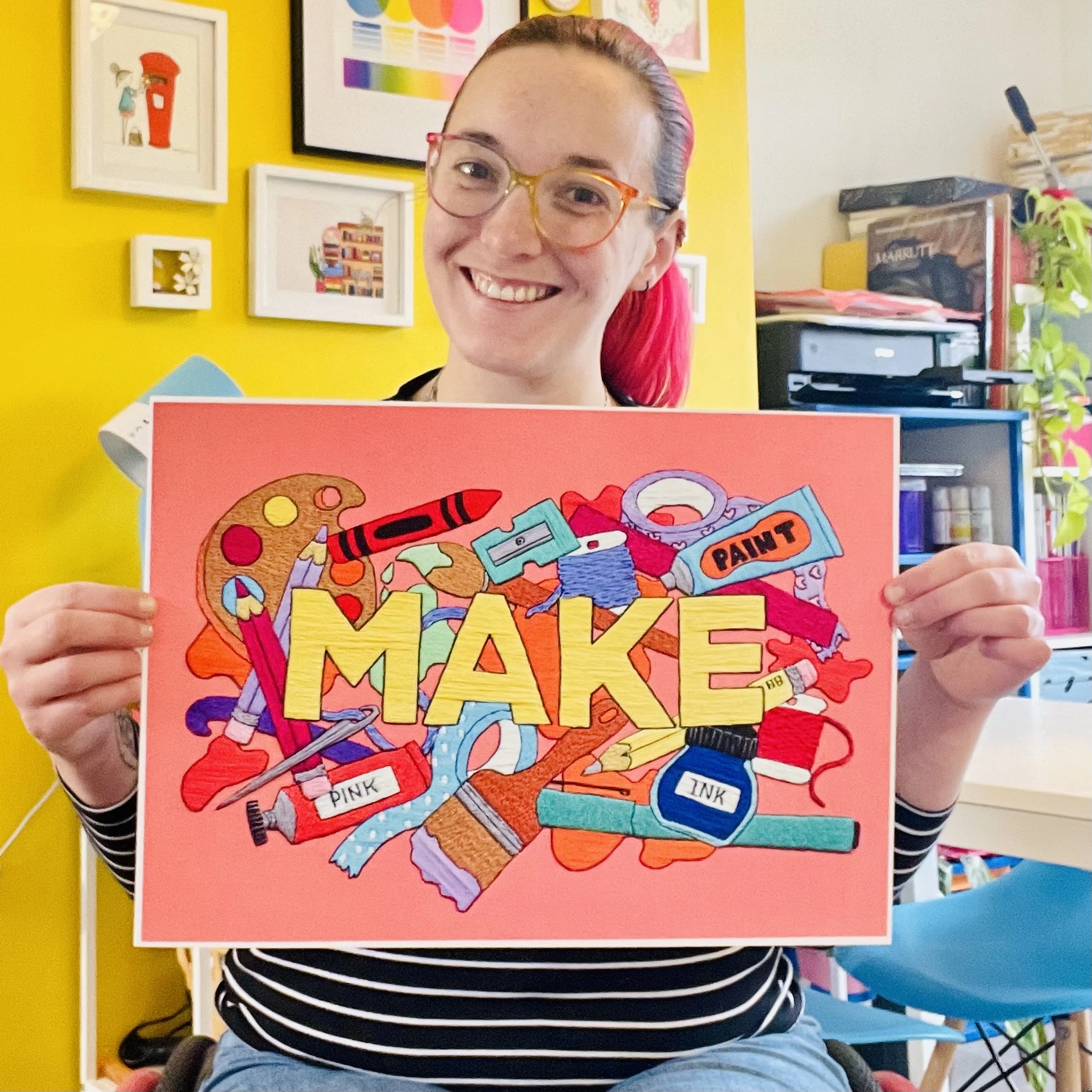 A white female with red hair and glasses holds a print of hand embroidered art that reads MAKE surrounded by illustrations of art and craft supplies