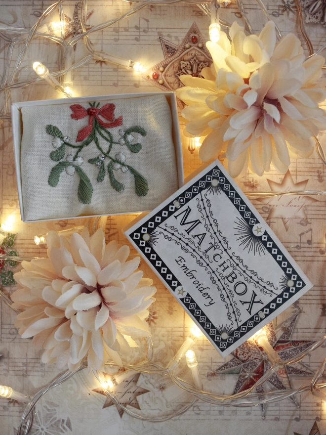 A sprig of mistletoe displayed in its matchbox.  surrounded by faux flowers and fairy lights.