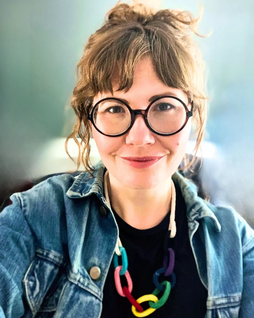 Portrait photo of This Thing is String founder Polly Benson, smiling to camera whilst wearing round glasses, a denim jacket, dark t-shirt and string bling necklace