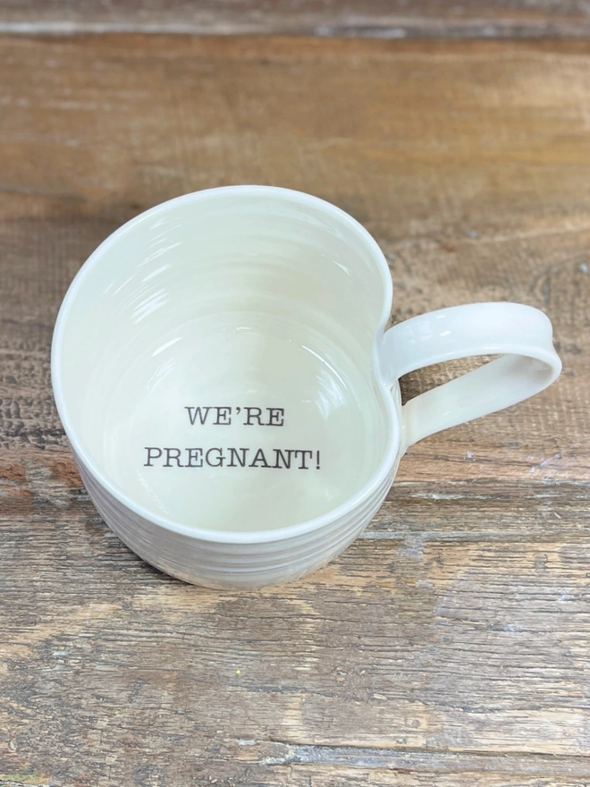 We're Pregnant! is announced in black text on the inside base of a hand thrown porcelain mug which sits atop a rustic table top