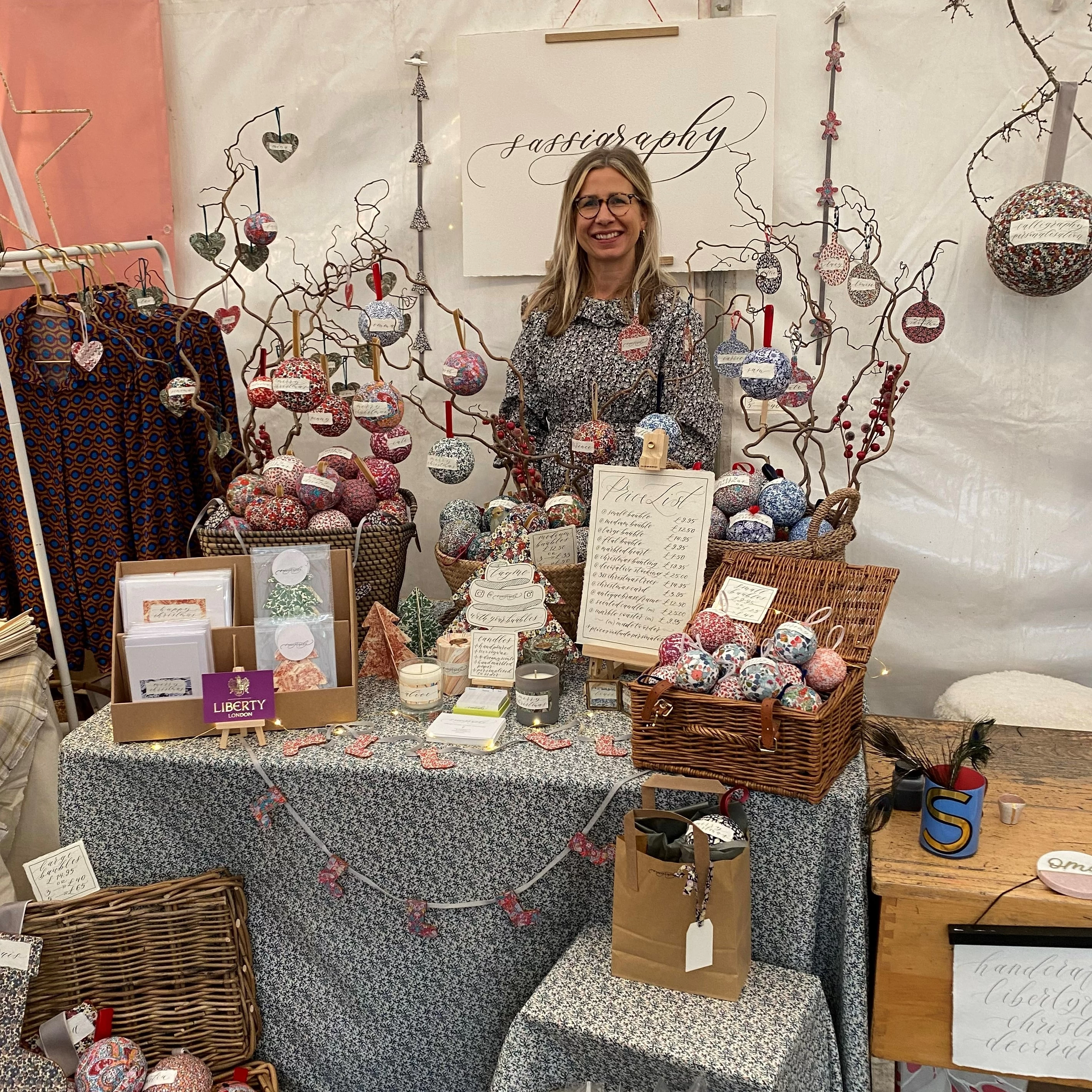 Sarah from Sassigraphy behind her stand at a Christmas fair selling personalised calligraphy Liberty fabric baubles and decorations. 