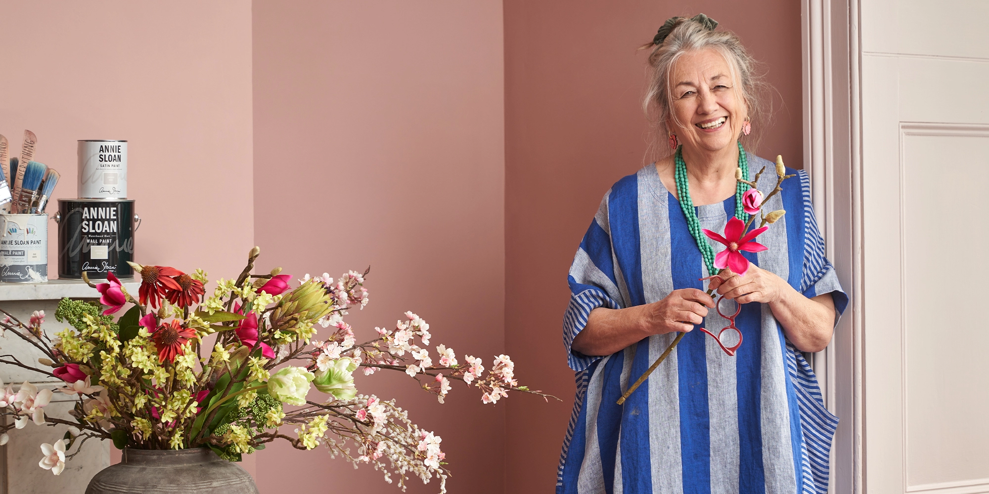 Annie Sloan CBE, founder of Annie Sloan, smiling at the camera wearing a blue striped dress, stood in front of a pink wall.