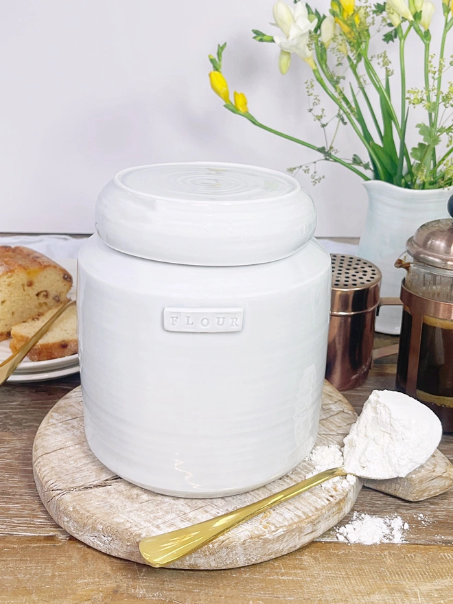 Flour hand thrown porcelain storage jar with lid. Sits on a rustic circular chopping board. Next to a gold spoon laden with flour. Jug of flowers, coffee pot and cake in the background.