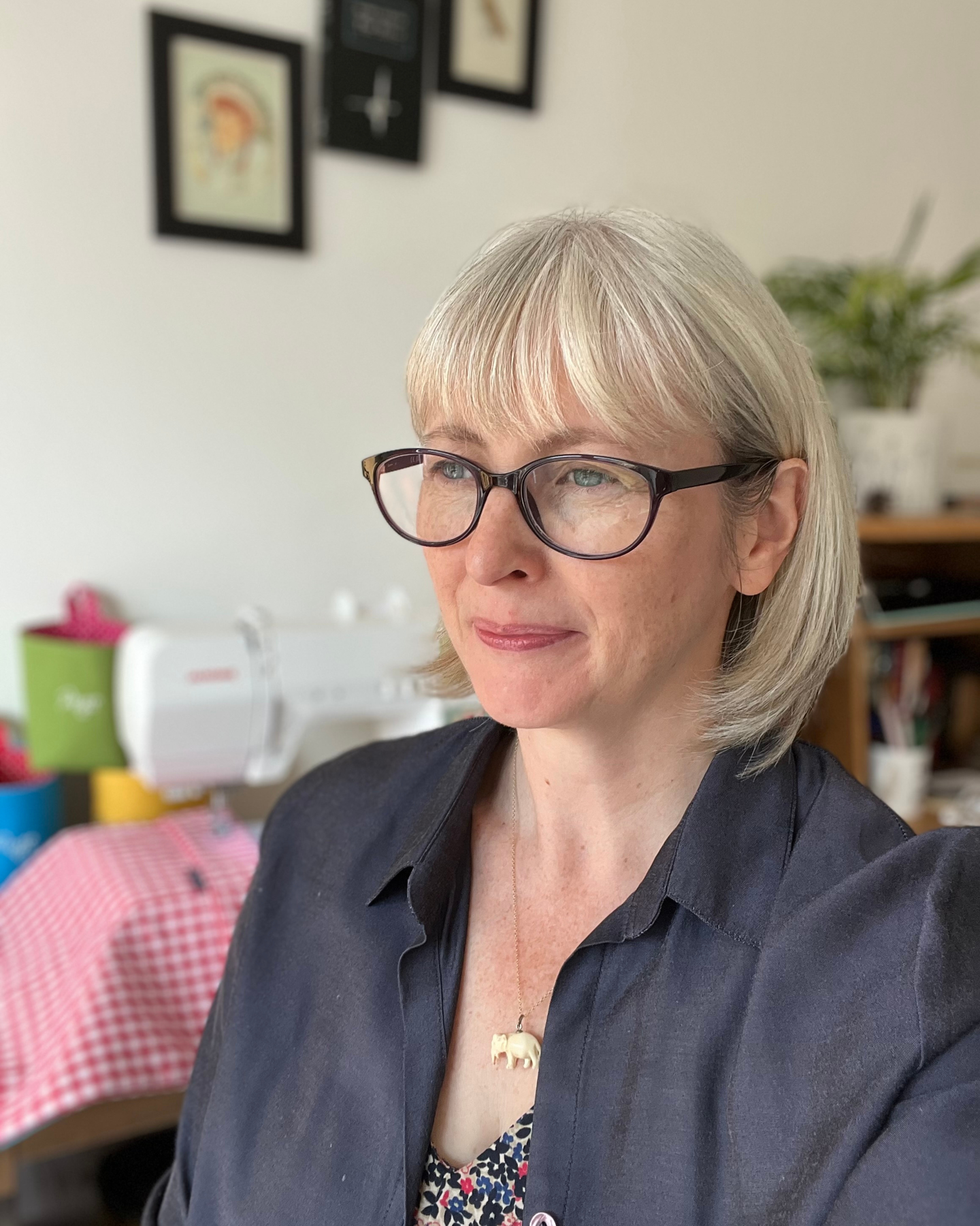 A photo of Sharon in her sewing nook with her sewing machine and colourful items she is creating in the background. Sharon has bobbed light hair, dark-rimmed glasses, and is wearing a deep blue blouse.