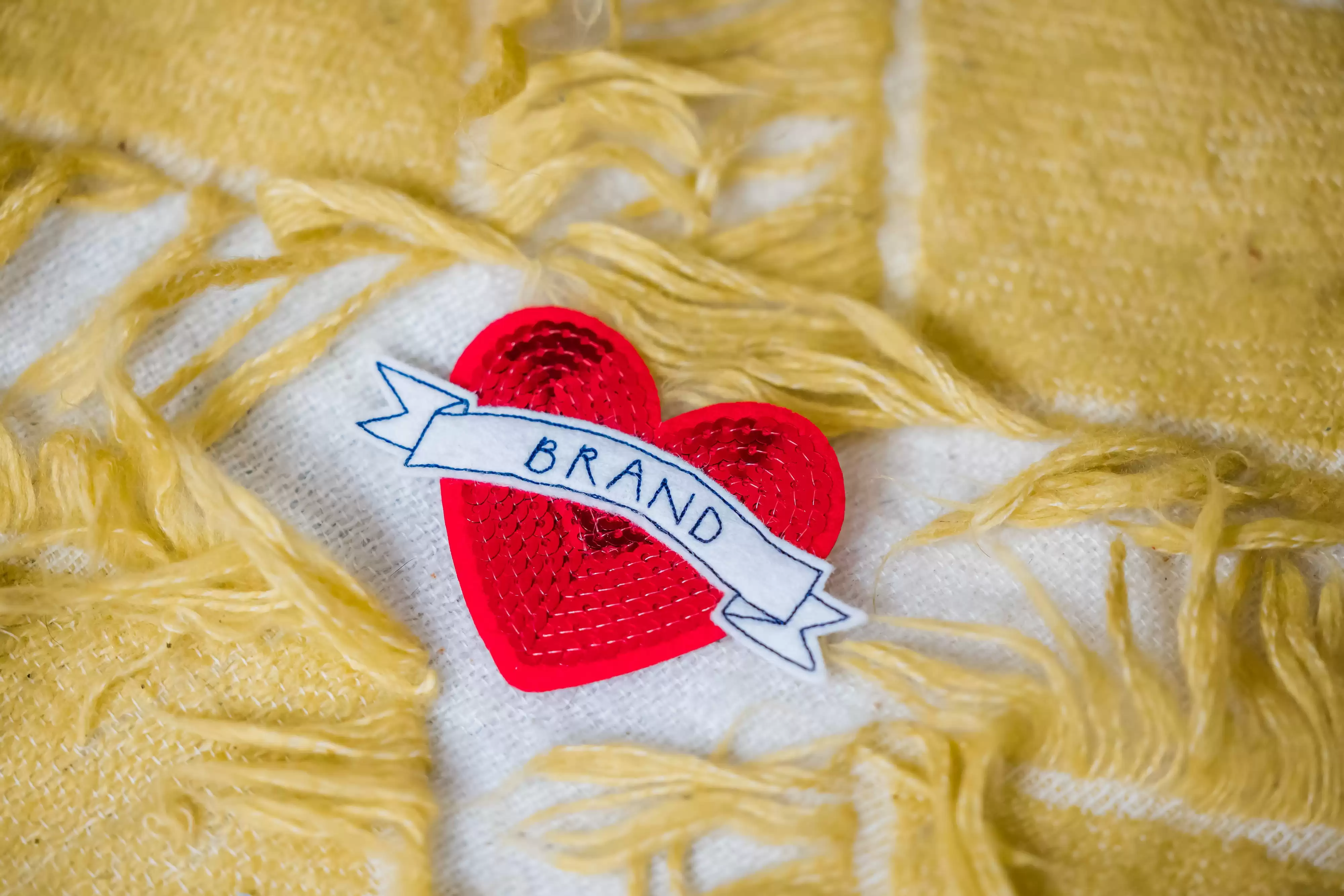 Red sequin heart with the word Brand across it on a yellow wool-background. 