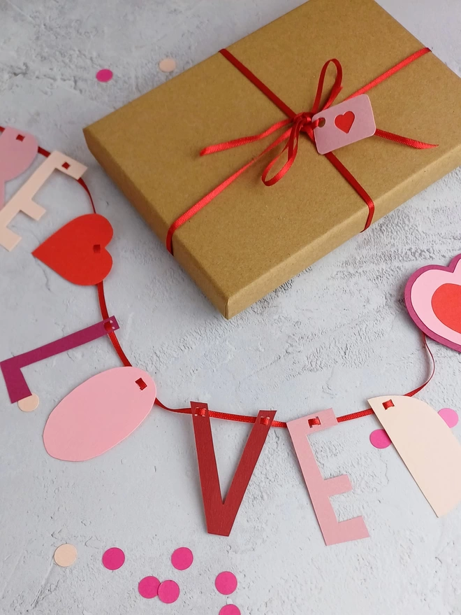 Gift wrap kraft coloured box tied with red ribbon and a small pink label with red heart. Below the box is part of the garland spelling the word love