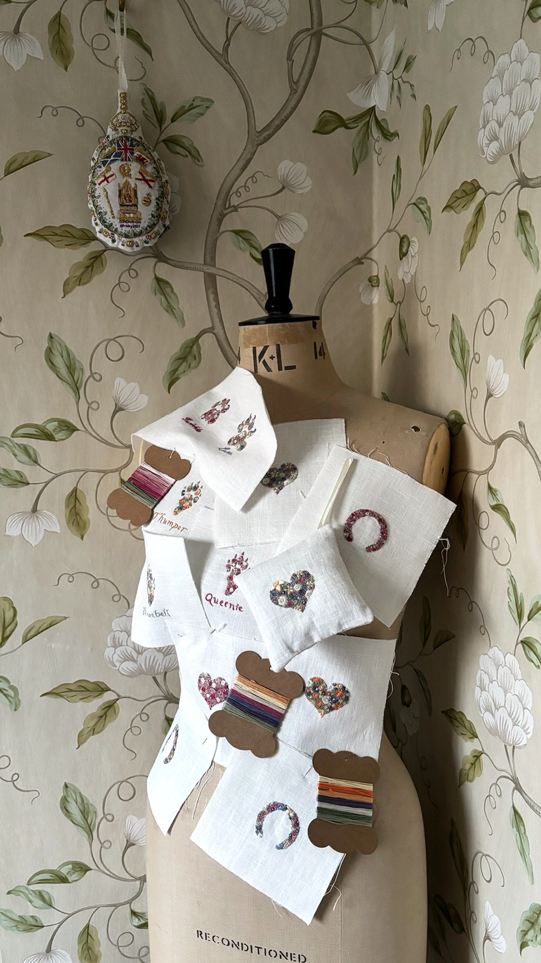 Embroidery designs and thread cards pinned to a tailors dummy in the corner of a botanical wallpapered room.  A coronation embroidery on a display hook in the background.