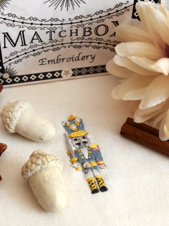 A mini embroidered nutcracker,  embroider in red or blue - colours are interchangeable. Displayed with decorative acorns and flora.