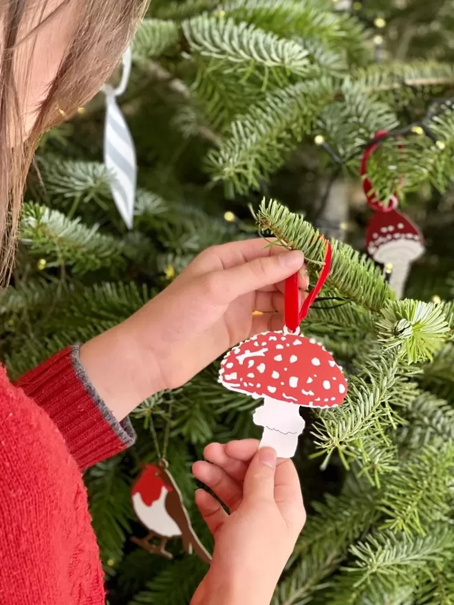 A girl wearing a red jumper is placing a red and white mushroom paper decoration onto a Christmas tree.  The mushroom decoration is threaded with bright red ribbon.