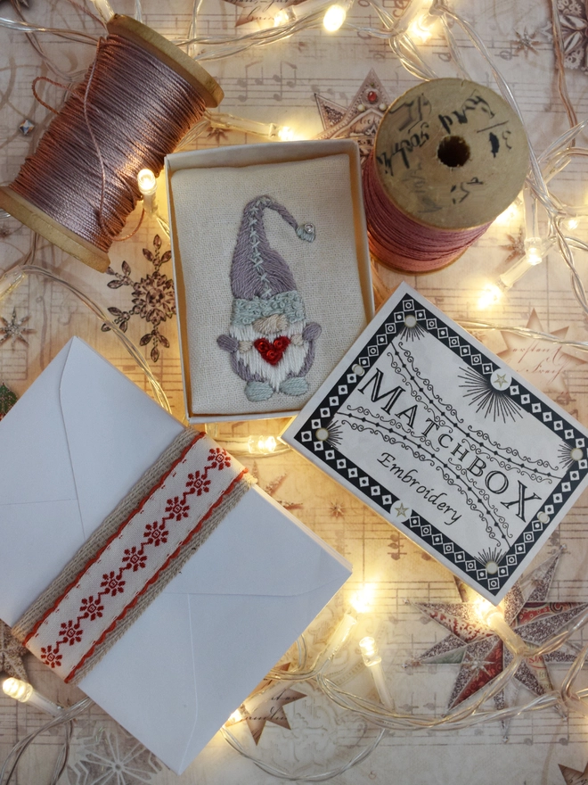 The embroidered Gonk displayed in its matchbox, surrounded by cotton reels, ribbon envelopes and fairy lights.