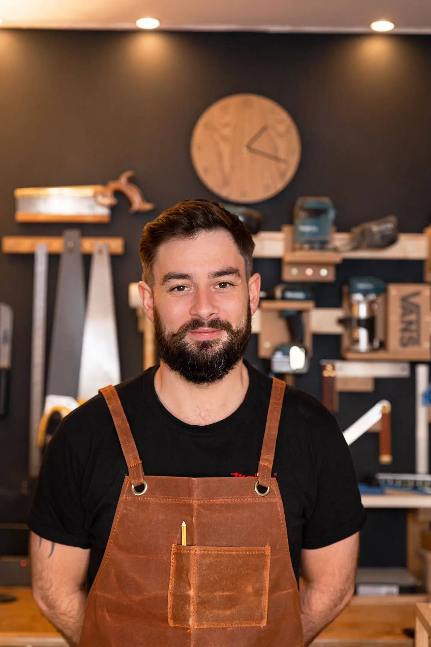 Shane wearing a black shirt and brown apron, smiling while standing in his workshop