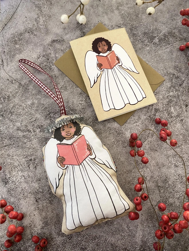 a Christmas angel plush facric decoration showing an afro Caribbean girl angel holding a song book laid next to a matching greeting card