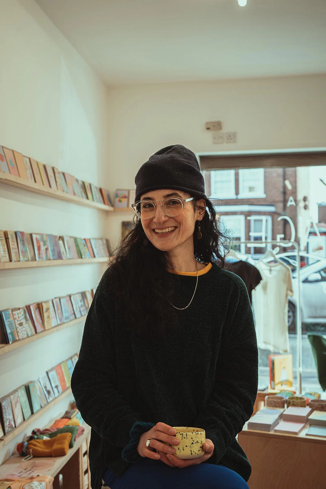 Nicola sits holding a coffee in her South Manchester gift shop. To her right a wall full of her greeting card designs, behind her a display of the stationery she designs. 