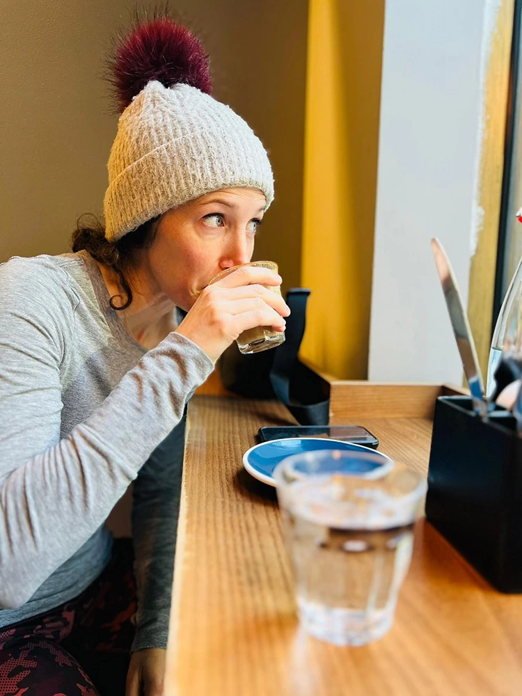 An image of Chloe wearing a woolly hat, drinking coffee and looking out of the window