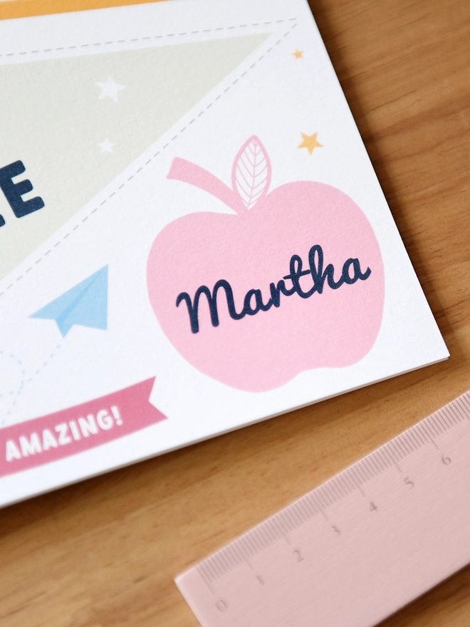  A "1st Day of Reception" card lies on a wooden desk surrounded by school supplies like scissors, pencils, a ruler, and a pencil case. The card includes a personalised name, encouraging messages, and playful designs.