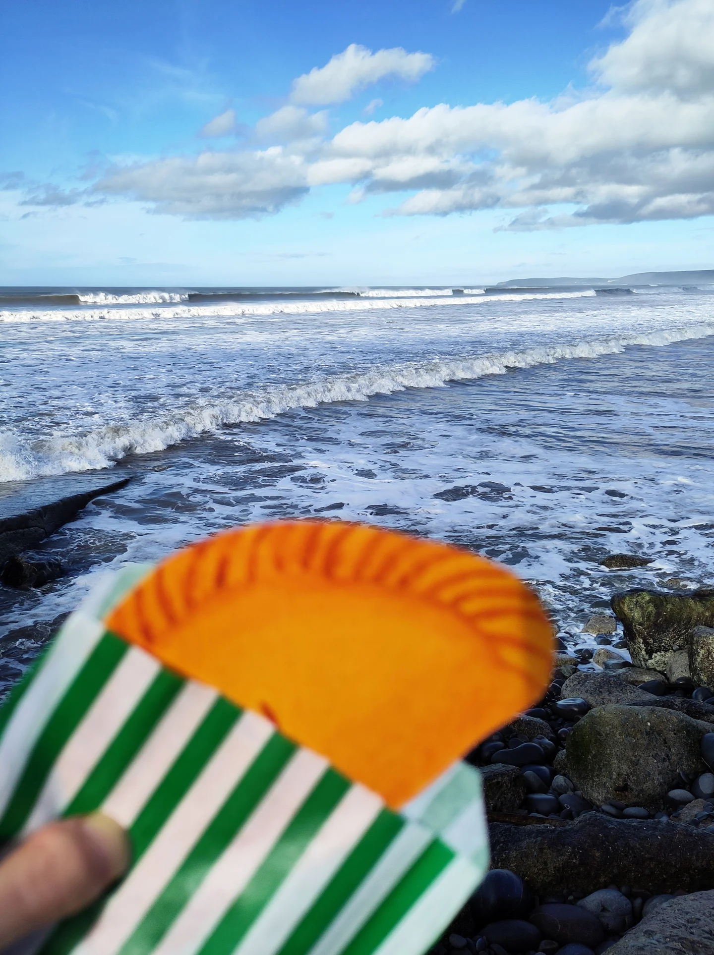 a hand-painted pasty held up in front of an ocean view as if you're about to take a bite out of it