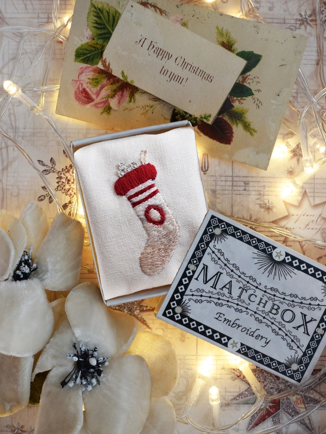 An embroidered stocking displayed in its matchbox, with fairy lights and Christmas decorations.